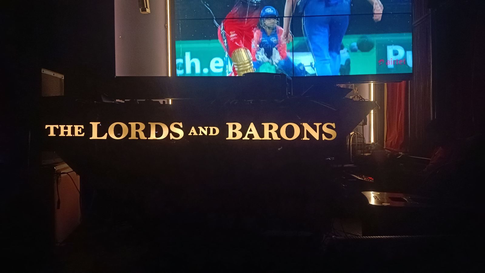 Illuminated gold The Lords and Barons signage beneath a live cricket screening